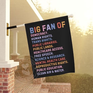 May include: A black flag with the text "BIG FAN OF" in pink, blue, and orange. The flag lists support for democracy, human rights, public libraries, and more. The flag is hanging outside a building.