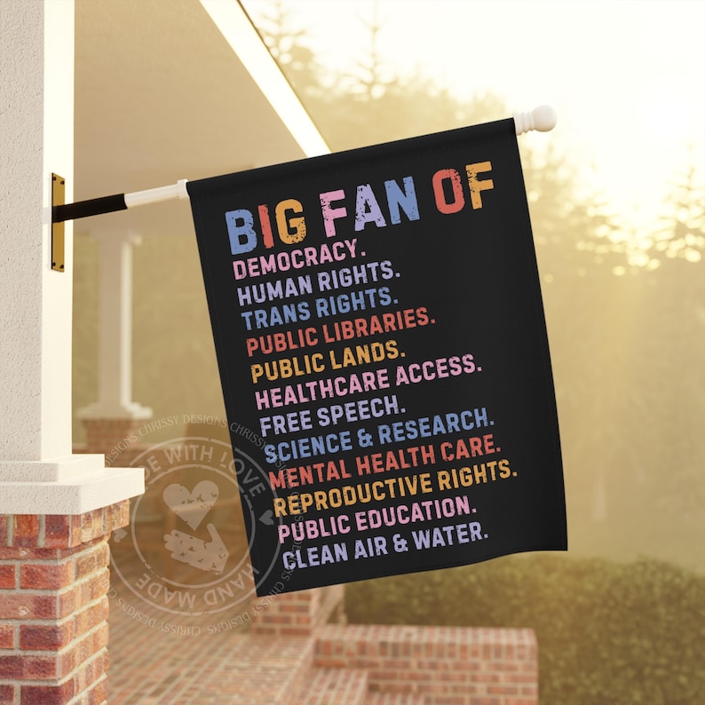 May include: A black flag with the text "BIG FAN OF" in pink, blue, and orange. The flag lists support for democracy, human rights, public libraries, and more. The flag is hanging outside a building.