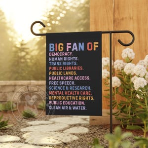 May include: A black garden flag with the text "BIG FAN OF" in blue, orange, and pink. Below, a list of values including "DEMOCRACY," "HUMAN RIGHTS," and "TRANS RIGHTS" is displayed in white. The flag is displayed on a black metal stand.