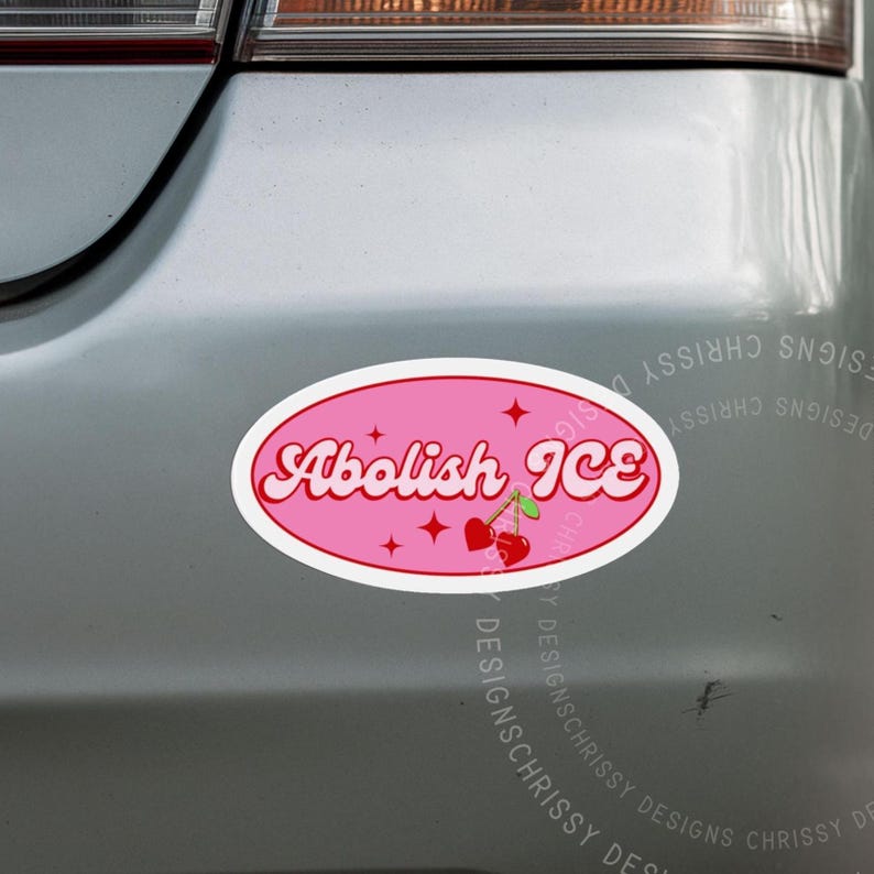 May include: A pink oval bumper sticker with the text "Abolish ICE" in a retro font. The sticker features cherries and star accents. The sticker is displayed on a silver car bumper.