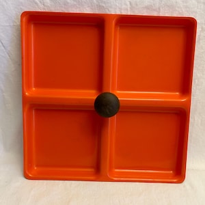 May include: A square, orange serving tray with four compartments. A dark brown, round handle is centered on the tray. The tray is made of a glossy material and is likely used for serving snacks or appetizers.
