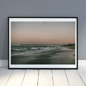 May include: A framed photograph of a beach scene with a wooden pier extending into the ocean. The water is a dark green color and the sky is a light pink and orange color. The photo is framed in a black frame.