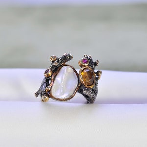 Puede incluir: Un anillo de plata único con una gran perla blanca irregular y una piedra más pequeña de color ámbar. Acentuado con pequeñas piedras preciosas rojas y detalles metálicos dorados, el anillo tiene un diseño texturizado y orgánico.