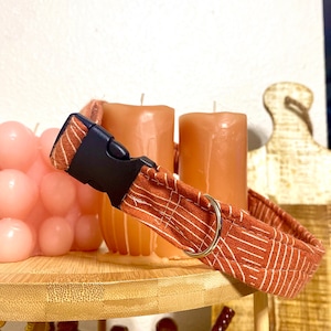 May include: A brown and white striped dog collar with a black buckle and a silver D-ring. The collar is on a wooden surface with candles in the background.