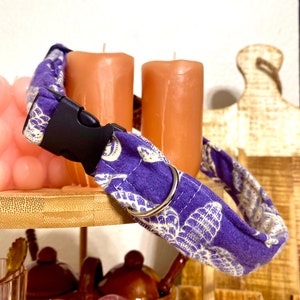 May include: Purple dog collar with a white butterfly pattern and a silver buckle. The collar is on a wooden surface with candles and a pink heart-shaped stone.
