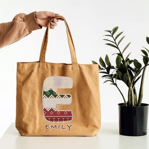 May include: A brown canvas tote bag with a red, green, and white patterned letter "E" and the name "Emily" written below.