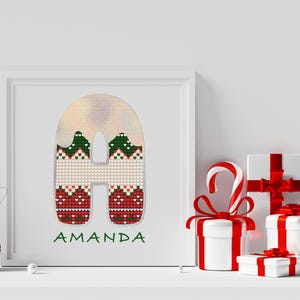 May include: A white frame with a red, green, and white knitted letter "A" with the name "Amanda" written below. The frame is on a white shelf with wrapped gifts and a candy cane.