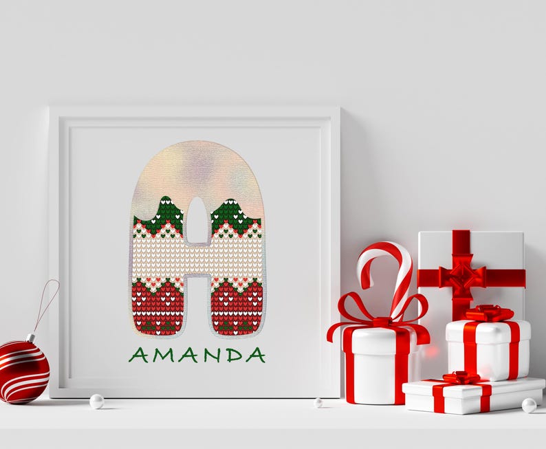 May include: A white frame with a red, green, and white knitted letter "A" with the name "Amanda" written below. The frame is on a white shelf with wrapped gifts and a candy cane.