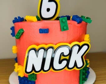 May include: A red and white Lego-themed cake with a number 6 on top and the name "NICK" on the side. The cake is decorated with colourful Lego bricks.