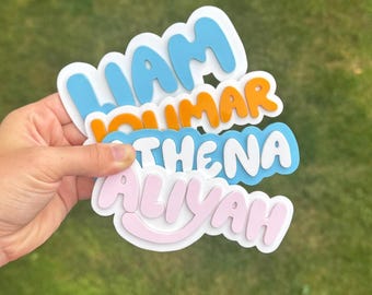 May include: A stack of four custom name stickers with a white border. The top sticker is light blue with the name LIAM, the second is orange with JOUMAR, the third is light blue with ATHENA, and the bottom sticker is light pink with ALIYAH.