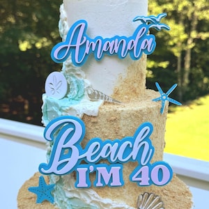 May include: A three-tiered cake decorated with a beach theme. The cake is adorned with blue and white frosting, edible sand, and sea-themed decorations. The cake topper reads "Amanda Beach I'm 40".