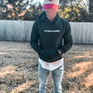 May include: A person wearing a black hoodie with the text "Life is short, buy the sneakers." in white, blue jeans, and a white and red baseball cap with the Ohio State logo.