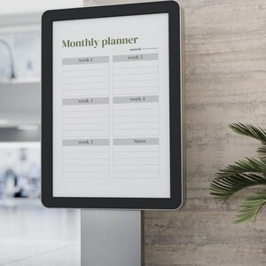 May include: A digital monthly planner with a black frame and a silver stand. The planner displays the text "Monthly planner" in green, with sections for weeks and notes. The background is a neutral wall.