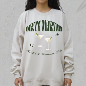 Puede incluir: Una sudadera de color beige claro con un estampado gr&aacute;fico verde de dos copas de martini con aceitunas y el texto "Dirty Martini Health & Wellness Club".