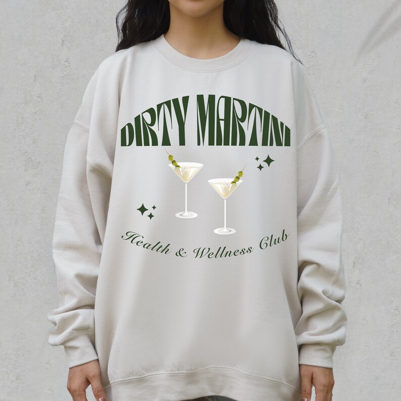 Puede incluir: Una sudadera de color beige claro con un estampado gr&aacute;fico verde de dos copas de martini con aceitunas y el texto "Dirty Martini Health & Wellness Club".