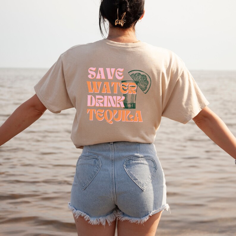 Pu&ograve; includere: Una persona indossa una maglietta beige con il testo "SAVE WATER DRINK TEQUILA" in lettere rosa e arancioni. La maglietta presenta un'illustrazione verde e nera di un bicchierino con una fetta di lime. La persona indossa pantaloncini di jeans blu con tasche a forma di cuore.