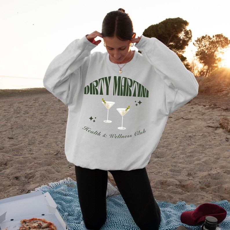 Puede incluir: Sudadera blanca con un gr&aacute;fico verde de dos copas de martini y el texto "Dirty Martini Health & Wellness Club".