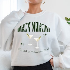 Puede incluir: Sudadera blanca con un gr&aacute;fico verde que dice "Dirty Martini Health & Wellness Club" con dos copas de martini con aceitunas.
