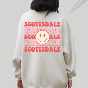May include: A beige sweatshirt with a pink and red graphic design on the back. The design features the word "Scottsdale" repeated four times with a smiley face in the center.