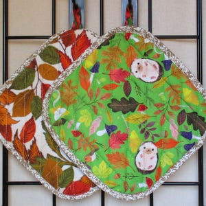 Könnte beinhalten: Zwei gesteppte Topflappen mit Herbstmotiven. Einer hat einen weißen Hintergrund mit orangefarbenen, roten und grünen Blättern. Der andere hat einen grünen Hintergrund mit Blättern, Pilzen und Igeln. Beide haben eine dekorative Bordüre.