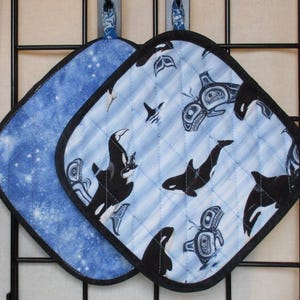 Può includere: Due presine quadrate trapuntate. Una è azzurra con un motivo a stelle bianche. L'altra è azzurra con un motivo di orche bianche e nere. Entrambe hanno bordi neri e anelli per appenderle.