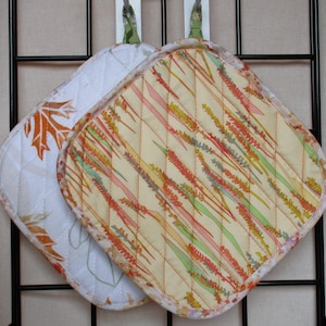 May include: Two quilted pot holders, one with a white background and a brown leaf design, and the other with a cream background and a colourful abstract design.