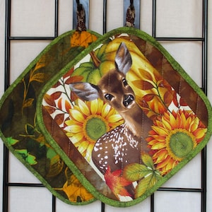 May include: Two quilted fabric pot holders featuring a printed design of a brown fawn with white spots, surrounded by yellow sunflowers, green pumpkin foliage, and autumn leaves. Each pot holder has a green fabric border and a hanging loop.