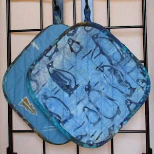 Puede incluir: Dos agarraderas de olla acolchadas azules con un patrón de pingüinos. Las agarraderas tienen un fondo azul claro con pingüinos azul oscuro y un contorno blanco. Las agarraderas tienen un lazo para colgar.