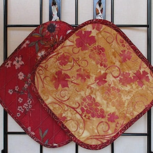May include: Two square quilted fabric pot holders with rounded corners. One features a red background with a floral pattern, while the other has a tan background with a red grape and vine motif. Both have a dark red fabric border and a hanging loop.
