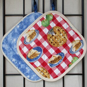 Peut inclure: Deux maniques en tissu matelassé avec des boucles de suspension. L'une présente un motif vichy rouge et blanc avec des parts de tarte aux pommes et des tartes entières. L'autre est bleue avec des étoiles blanches. Bords en tissu crème.