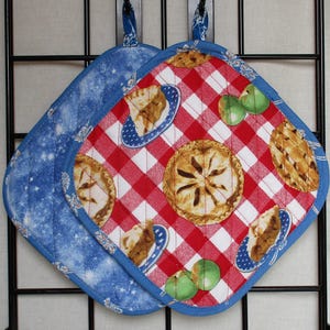 May include: Two quilted pot holders. One is blue with a white star pattern. The other has a red and white checkered pattern with images of apple pie slices and whole pies. Both have blue trim and hanging loops.