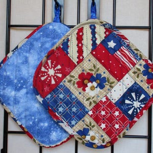 Puede incluir: Dos agarraderas acolchadas. Una es azul con un patrón de estrellas blancas. La otra es un diseño de patchwork con cuadrados rojos, blancos y azules con estrellas, rayas y motivos florales. Ambas tienen lazos azules para colgar.