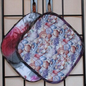 May include: Two quilted pot holders, one with a red and white quilted pattern and the other with a seashell pattern in shades of blue, purple, and brown.