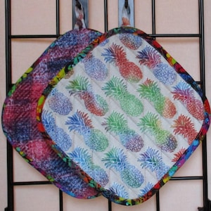 May include: Two colourful pot holders with a pineapple pattern. One pot holder is a dark blue, red and purple tie-dye pattern. The other pot holder is a white background with a blue and green pineapple pattern.