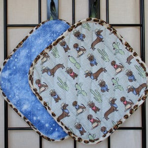 May include: Two quilted pot holders with rounded corners and a brown coffee bean patterned trim. One features a light blue fabric with a repeating pattern of dachshunds wearing cowboy hats and bandanas alongside green cacti. The other is blue.