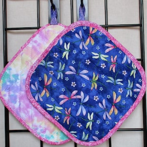 Puede incluir: Dos agarraderas de olla acolchadas con ribete rosa. Una es de tela azul con un patrón de libélula y la otra es un patrón tie-dye en rosa, morado y blanco.