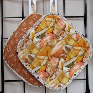 Può includere: Due presine da forno marroni e bianche con una texture trapuntata. Le presine presentano un motivo con diversi tipi di formaggio.