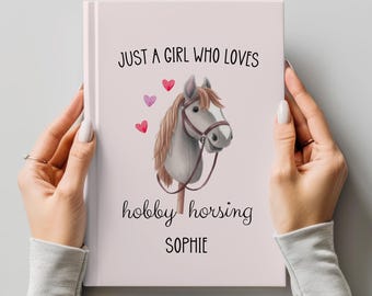 May include: A light pink journal with the text "JUST A GIRL WHO LOVES hobby horsing SOPHIE" and a watercolor illustration of a horse head with hearts. The journal is held by a person.