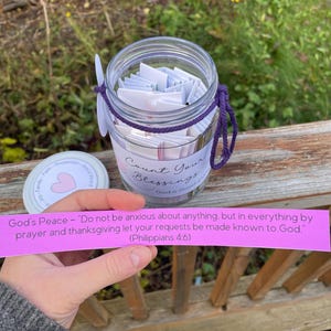 Op de afbeelding: Een glazen pot gevuld met kleine witte papiertjes, vastgebonden met een paars koord. Op de pot staat "Count Your Blessings". Een roze papieren strook met tekst wordt voor de pot gehouden.