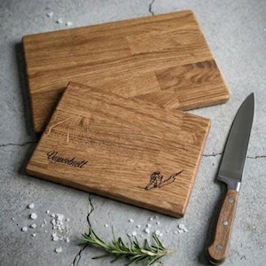 May include: Two rectangular wooden cutting boards with a light brown finish. One board has the word "Ungerbrett" and a skier etched into the wood. A stainless steel knife with a wooden handle and a sprig of rosemary are next to the boards.