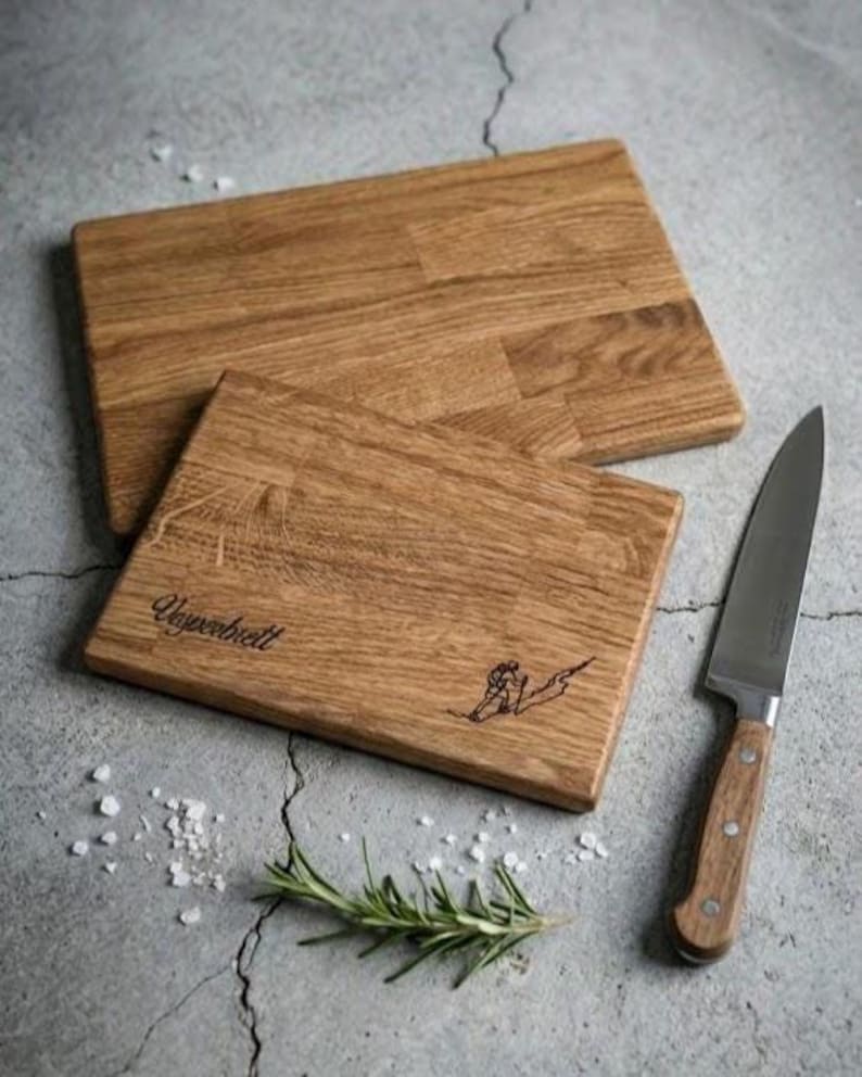 May include: Two rectangular wooden cutting boards with a light brown finish. One board has the word "Ungerbrett" and a skier etched into the wood. A stainless steel knife with a wooden handle and a sprig of rosemary are next to the boards.