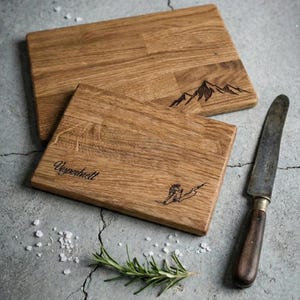 May include: Two rectangular wooden cutting boards with mountain and skier designs. One board has the word "Vesperbrett" etched on it. A knife, rosemary sprig, and salt are next to the boards.