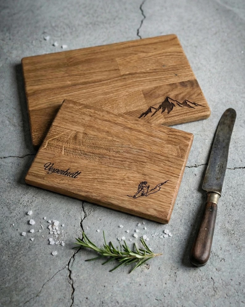 May include: Two rectangular wooden cutting boards with mountain and skier designs. One board has the word "Vesperbrett" etched on it. A knife, rosemary sprig, and salt are next to the boards.
