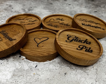 May include: A collection of oval wooden coasters with engraved text and heart designs. The coasters are made of light brown wood and feature phrases like "Lieblingsgast" and "Glücks Pils" in a dark font. The coasters are arranged on a textured gray surface.