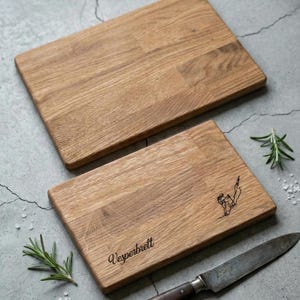May include: Two rectangular wooden cutting boards with a light brown finish. One board has the word "Vesperbrett" engraved, along with a small illustration. A knife and sprigs of rosemary are also present.