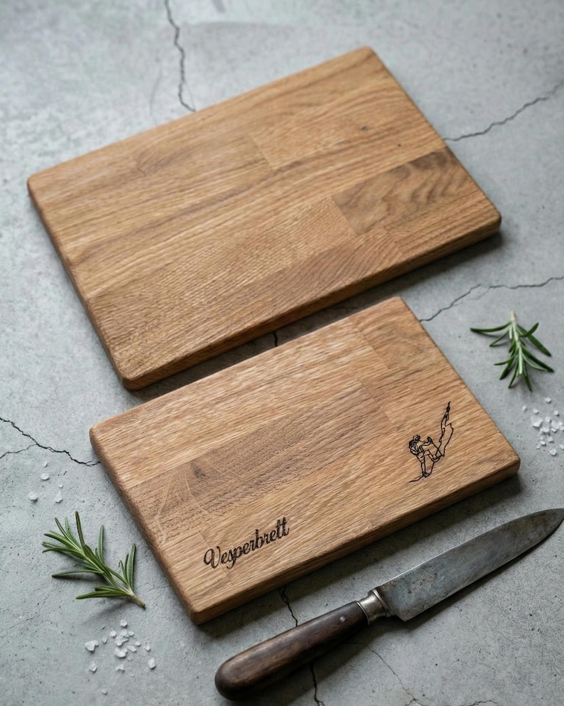 May include: Two rectangular wooden cutting boards with a light brown finish. One board has the word "Vesperbrett" engraved, along with a small illustration. A knife and sprigs of rosemary are also present.