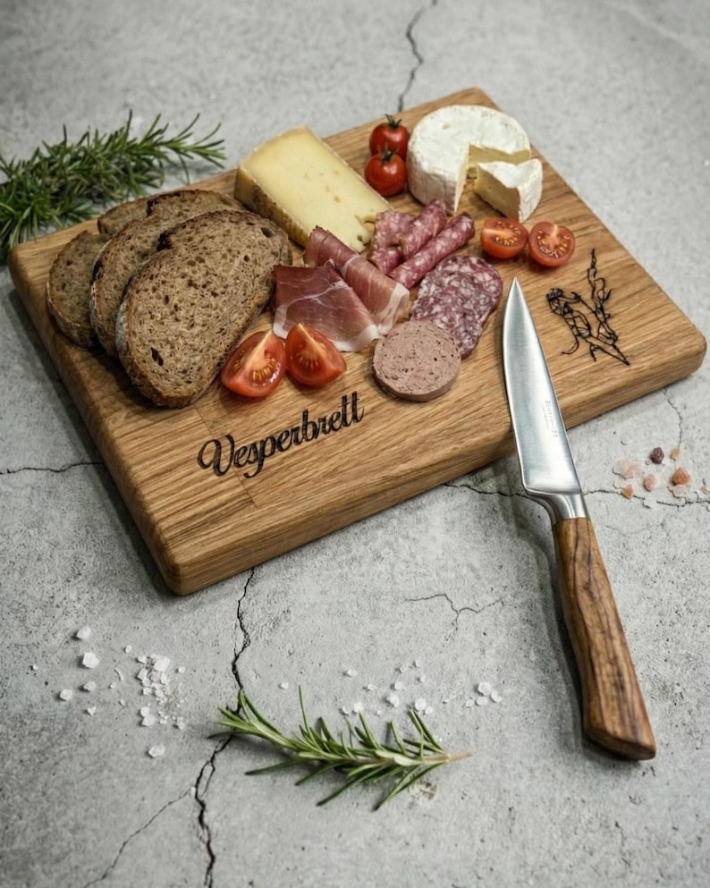 May include: A wooden charcuterie board with various cheeses, sliced meats, bread, and tomatoes. A stainless steel knife with a wooden handle rests on the board. The word "Vesperbrett" is etched on the board.