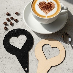 Könnte beinhalten: Eine Tasse Cappuccino mit einem Herzmuster im Schaum steht auf einer weißen Untertasse. Zwei herzförmige Untersetzer, einer schwarz und einer aus Naturholz, liegen neben der Tasse. Kaffeebohnen und ein Löffel sind ebenfalls zu sehen.