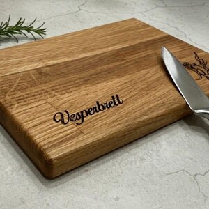 May include: A rectangular wooden cutting board with the word "Vesperbrett" engraved on it. A stainless steel knife rests on the board, next to a sprig of rosemary. The board has a natural wood grain.