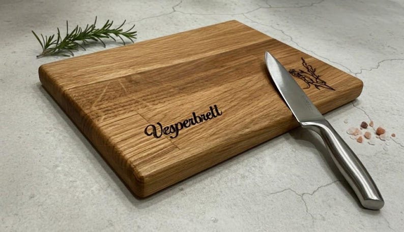 May include: A rectangular wooden cutting board with the word "Vesperbrett" engraved on it. A stainless steel knife rests on the board, next to a sprig of rosemary. The board has a natural wood grain.